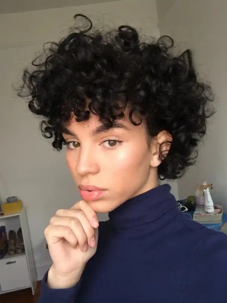 Woman with short curly pixie hairstyle wearing a navy blue turtleneck, posing indoors in natural light with soft makeup and glossy lips.